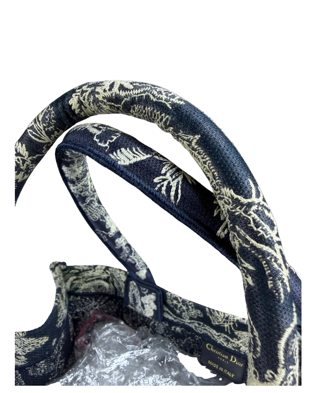 Close-up of a navy blue handbag handle with an intricate beige and white floral pattern, featuring a gold Christian Dior logo tag.