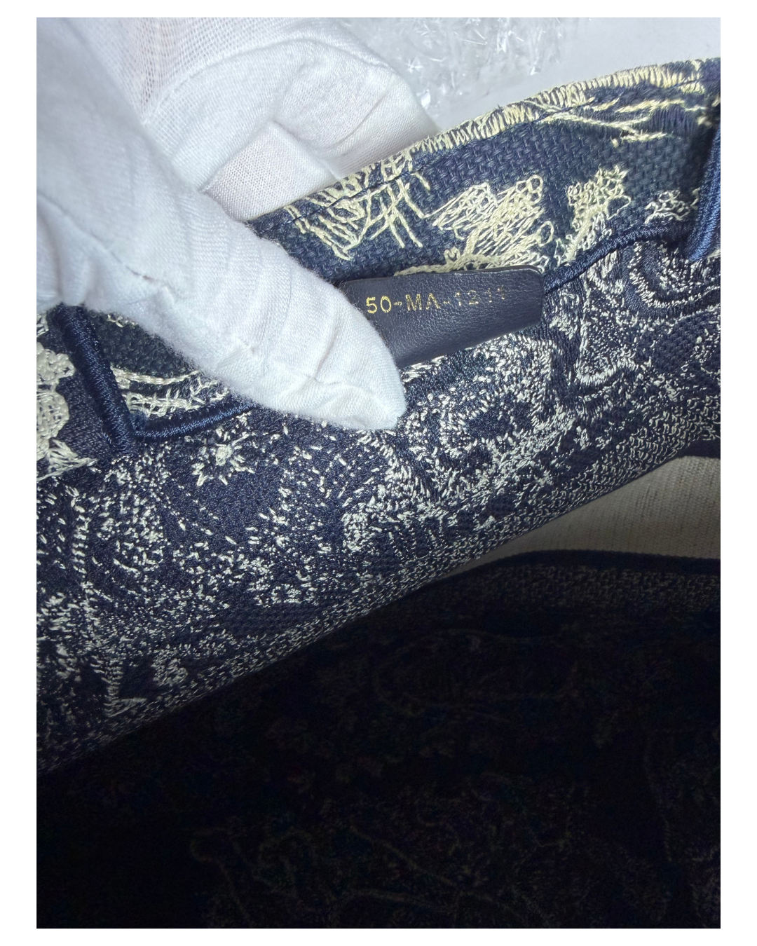 Close-up of a navy blue and beige patterned textile item being held by a white-gloved hand, with an interior size label visible reading 50-MA-1211.