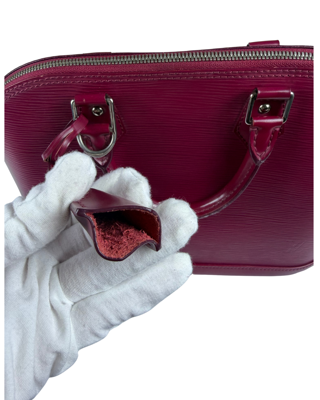 A close-up of a burgundy textured leather handbag with silver-tone hardware, shown with a white-gloved hand holding a key holder matching burgundy leather.