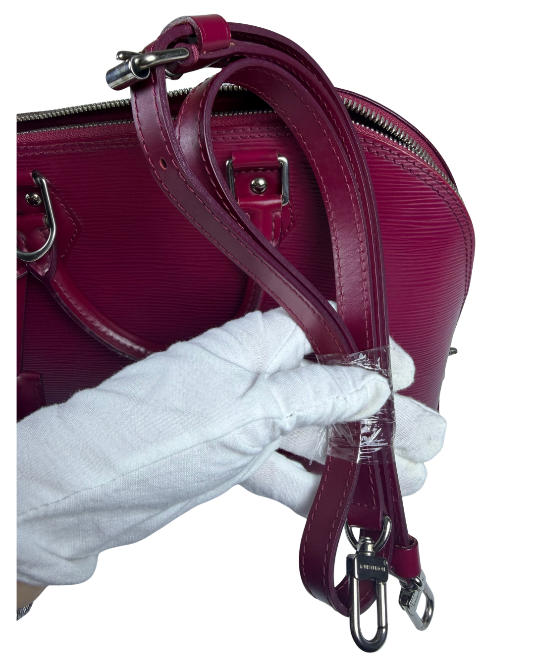 A close-up of a burgundy textured leather handbag with a white gloved hand holding its detachable shoulder strap, showing metal hardware and a clear protective wrap on the strap end.